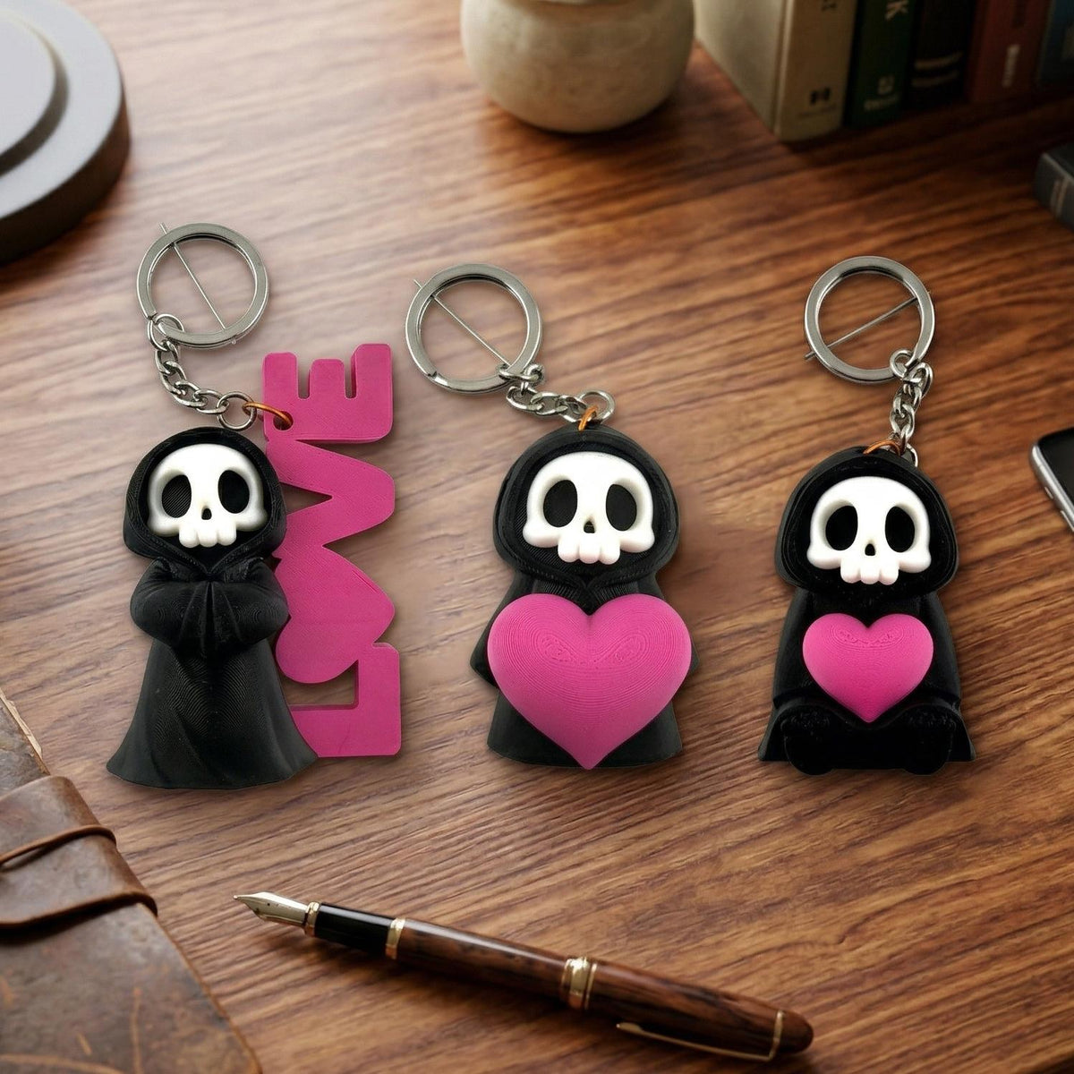 Three Love Reaper keyrings on a wooden desk: one with a pink LOVE sign, one holding a big heart, one sitting with a heart
