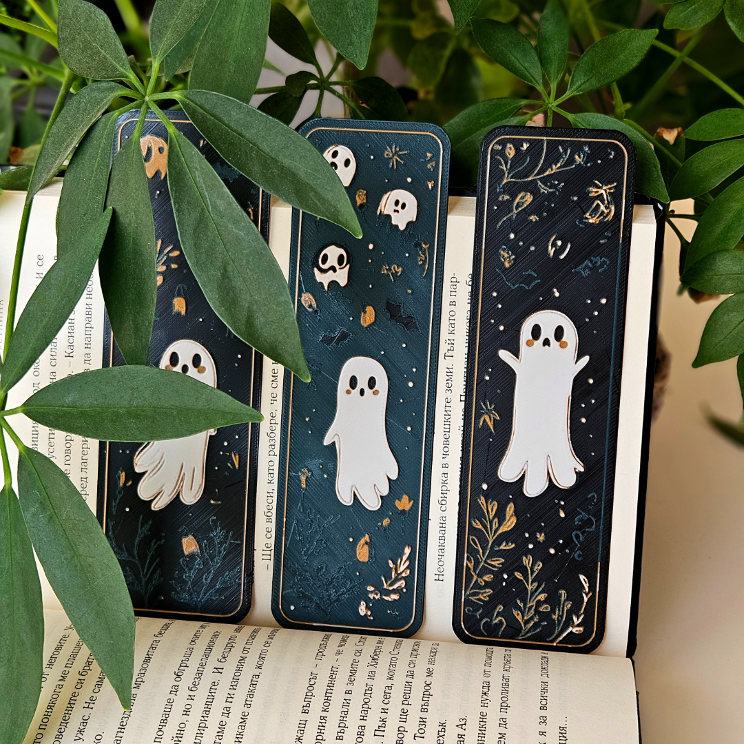 Three Halloween-themed bookmarks with ghost illustrations on an open book surrounded by green leaves.