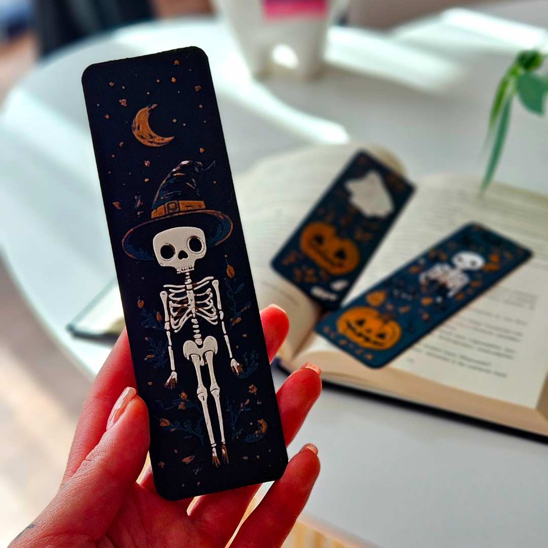 Halloween-themed bookmark with a skeleton design held by a hand, with more bookmarks and an open book in the background.