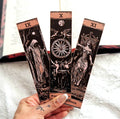 Three tarot card bookmarks held by a hand on a textured surface.