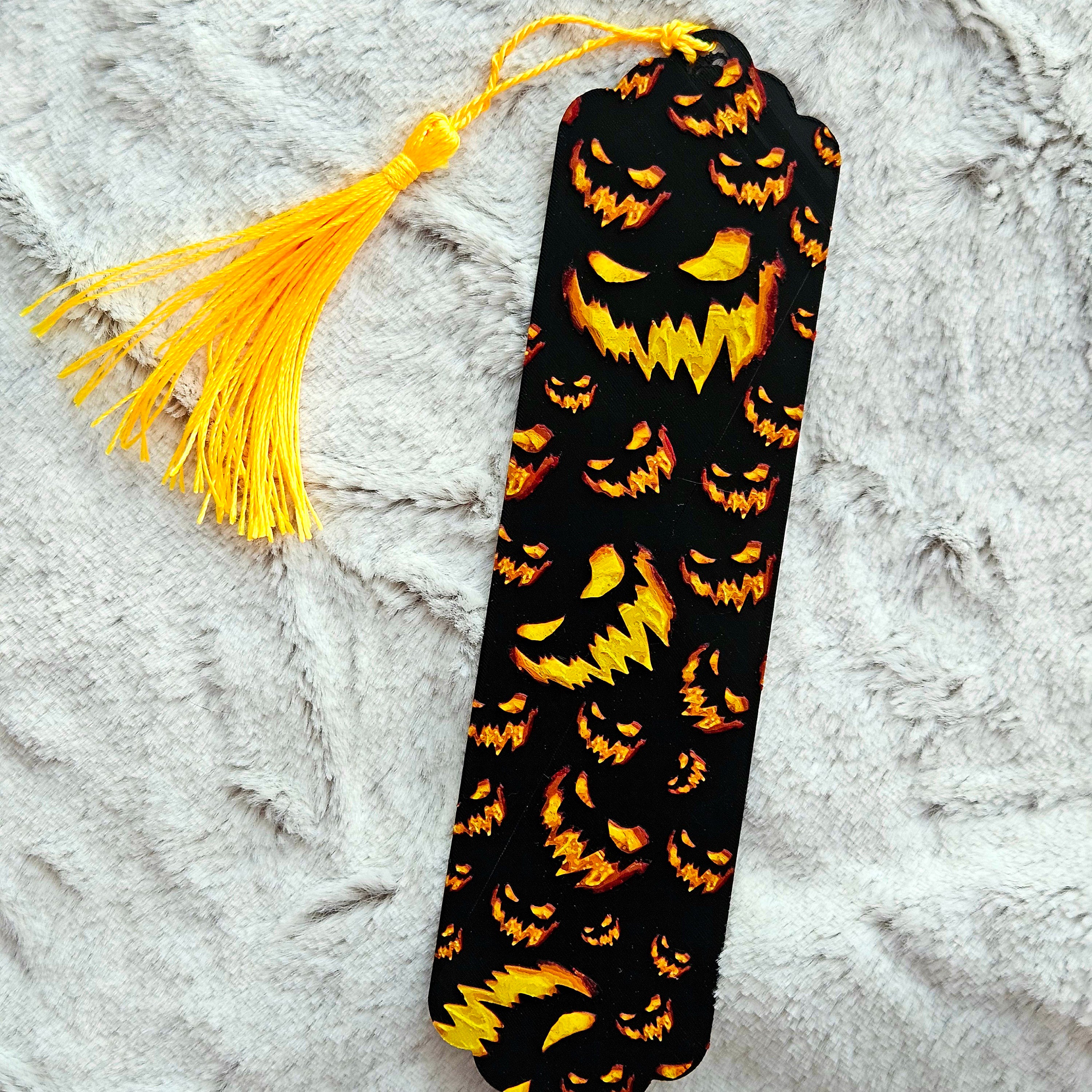 Black bookmark with orange jack-o'-lantern faces and a yellow tassel on a textured gray background