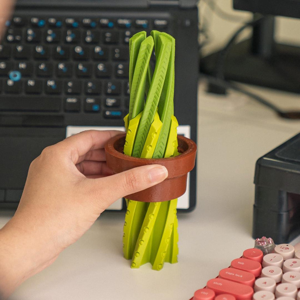 Hand holding a small pot with green folding tools on a desk with a laptop in the background