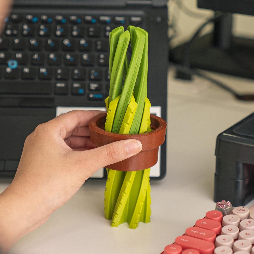 Hand holding a small pot with green folding tools on a desk with a laptop in the background