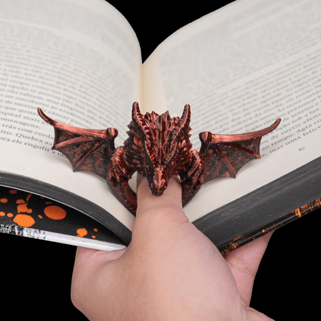 Copper dragon thumb page holder holding an open book flat, on a reader's thumb