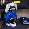 White dual controller stand holding a PS5 controller above a blue Xbox controller, beside a gaming monitor