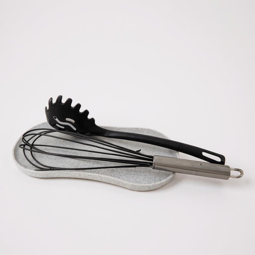 Black pasta server and whisk on a gray stone surface with a white background
