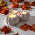 White ceramic pumpkin-shaped candle holders with lit candles on a white surface with autumn leaves.
