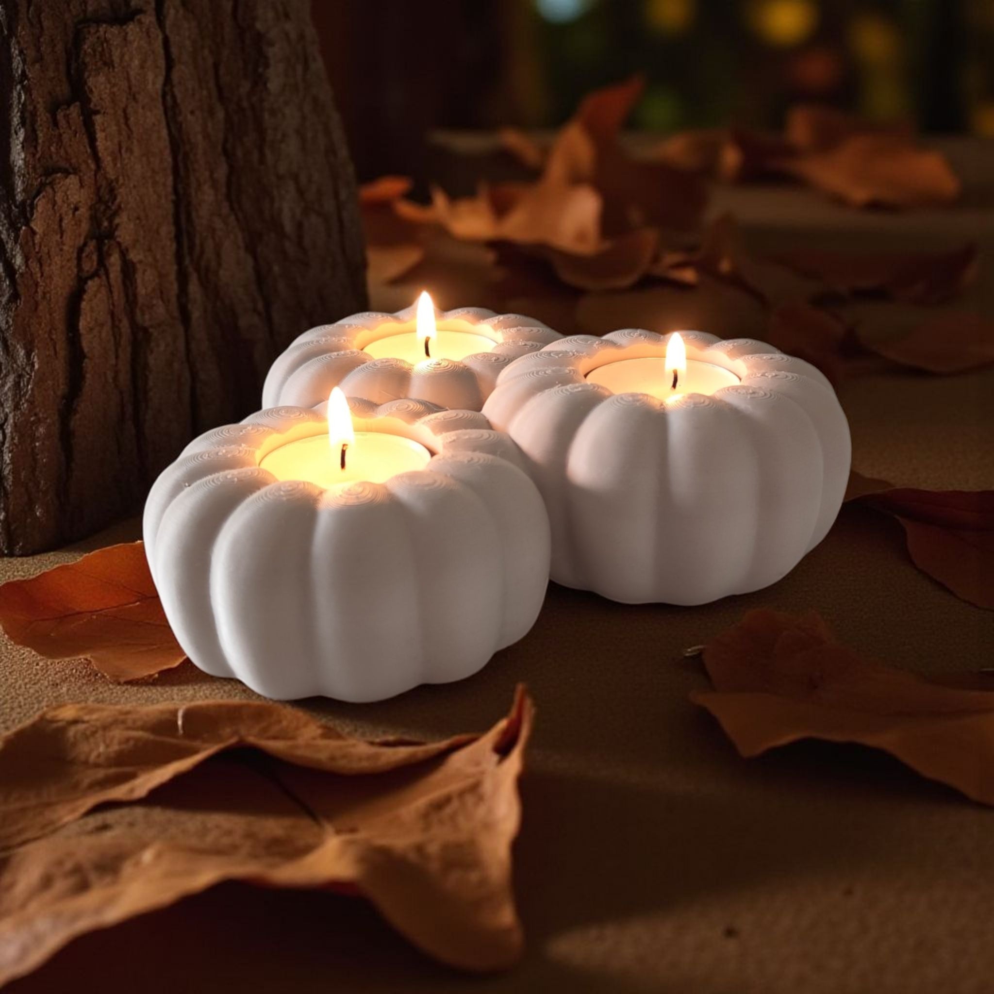 Three white pumpkin-shaped candles with lit flames on a surface with autumn leaves and a tree trunk in the background.