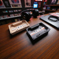 Two cassette tapes on a wooden surface with headphones in the background