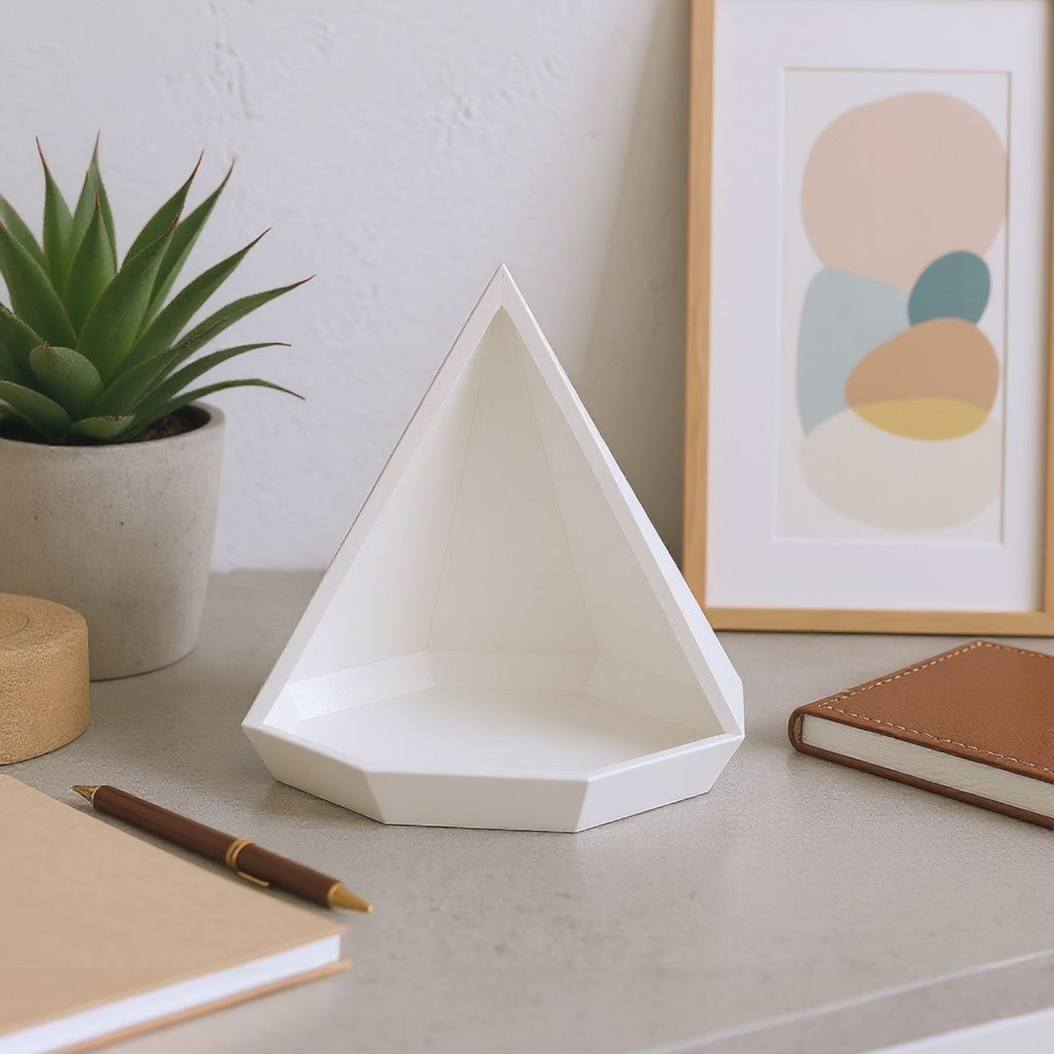 Geometric white vase on a desk with a plant, notebook, and pen in the background.