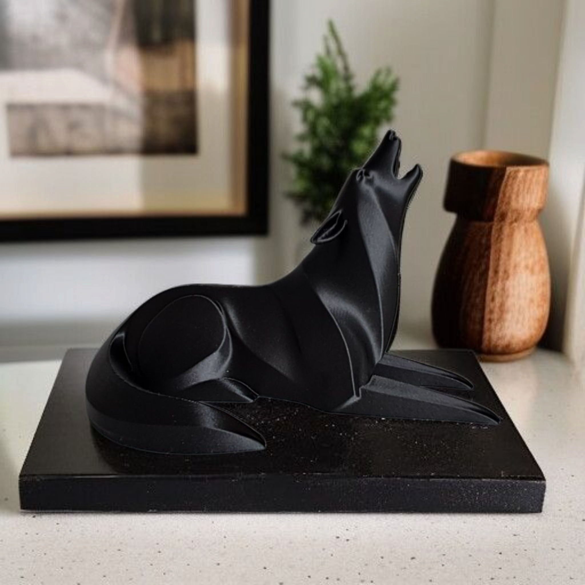 Black abstract sculpture on a white surface with a wooden vase and framed picture in the background.