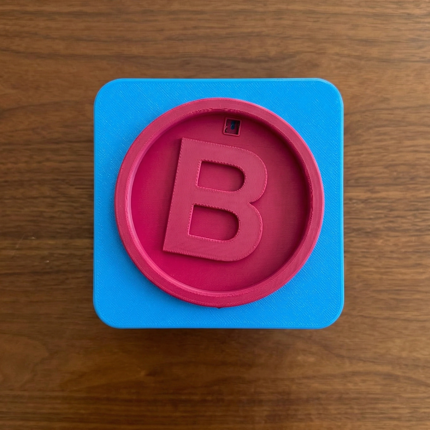 Blue square with a pink circle containing the letter 'B' on a wooden surface