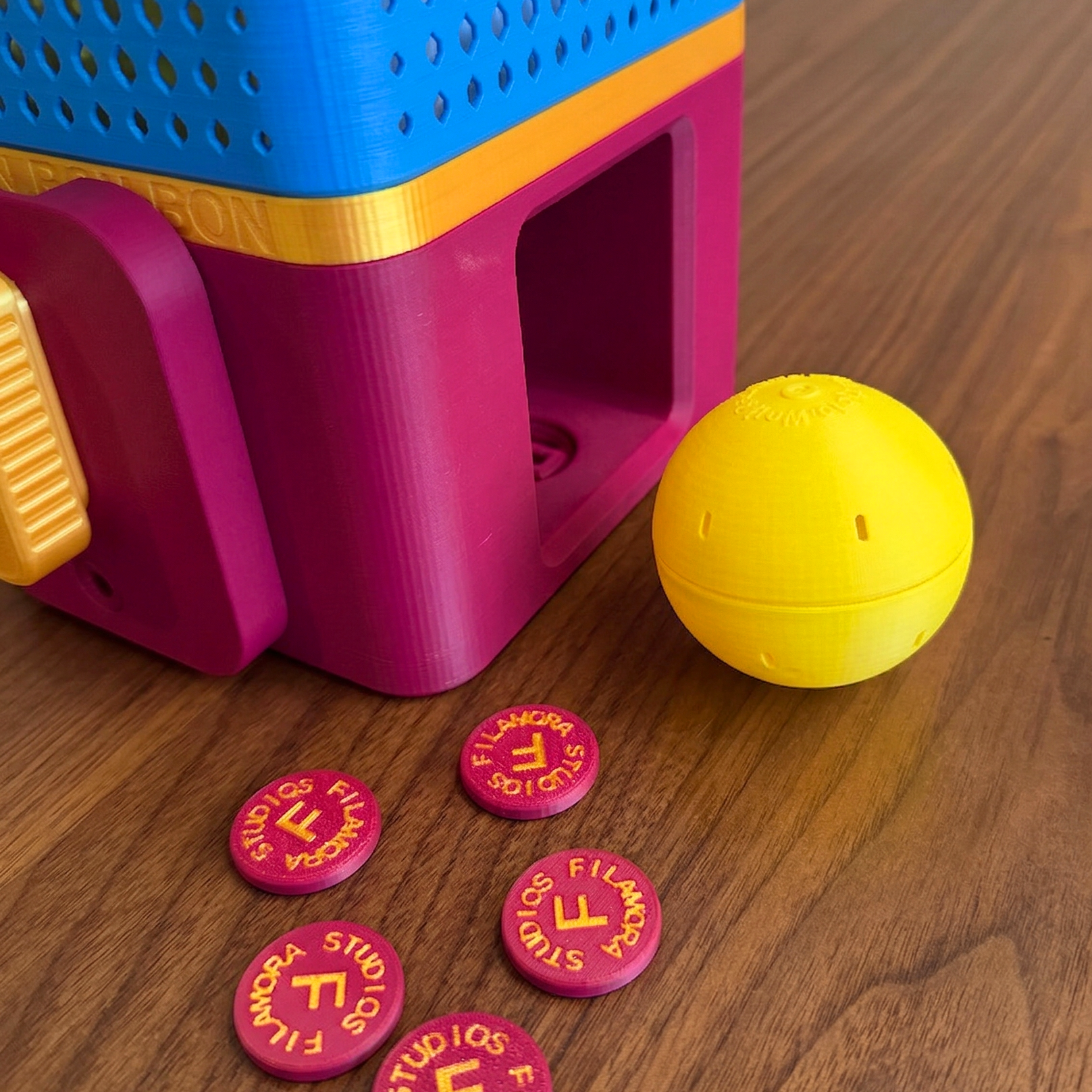 Colorful toy dispenser with a yellow ball and pink tokens on a wooden surface
