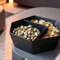 Black divided bowl with pistachios and almonds on a reflective surface.