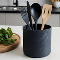 Black utensil holder with black cutlery and a wooden spatula on a kitchen counter.